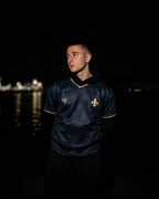Man wearing a dark blue sports jersey with a Bosnian lily, standing against a night waterfront background.

