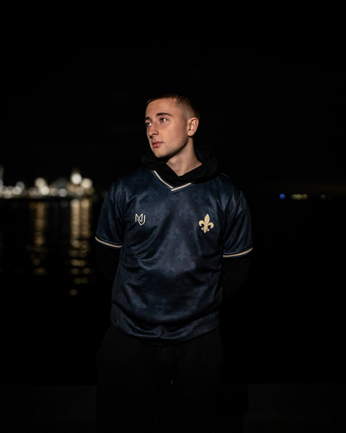 Man wearing a dark blue sports jersey with a Bosnian lily, standing against a night waterfront background.
