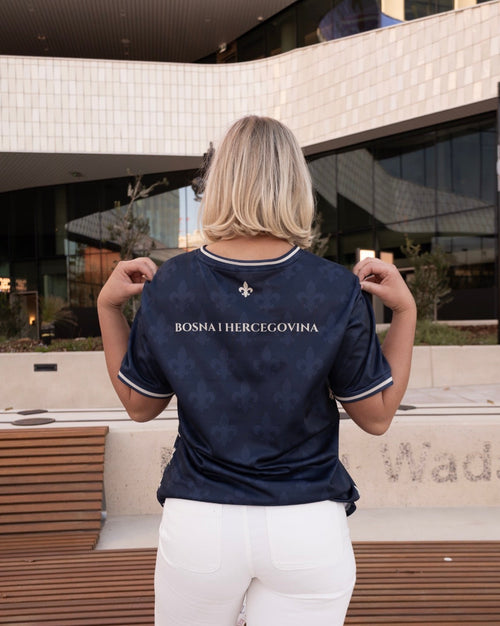 Person wearing a navy blue Bosnian soccer jersey styled with white jeans, standing in front of a modern building.